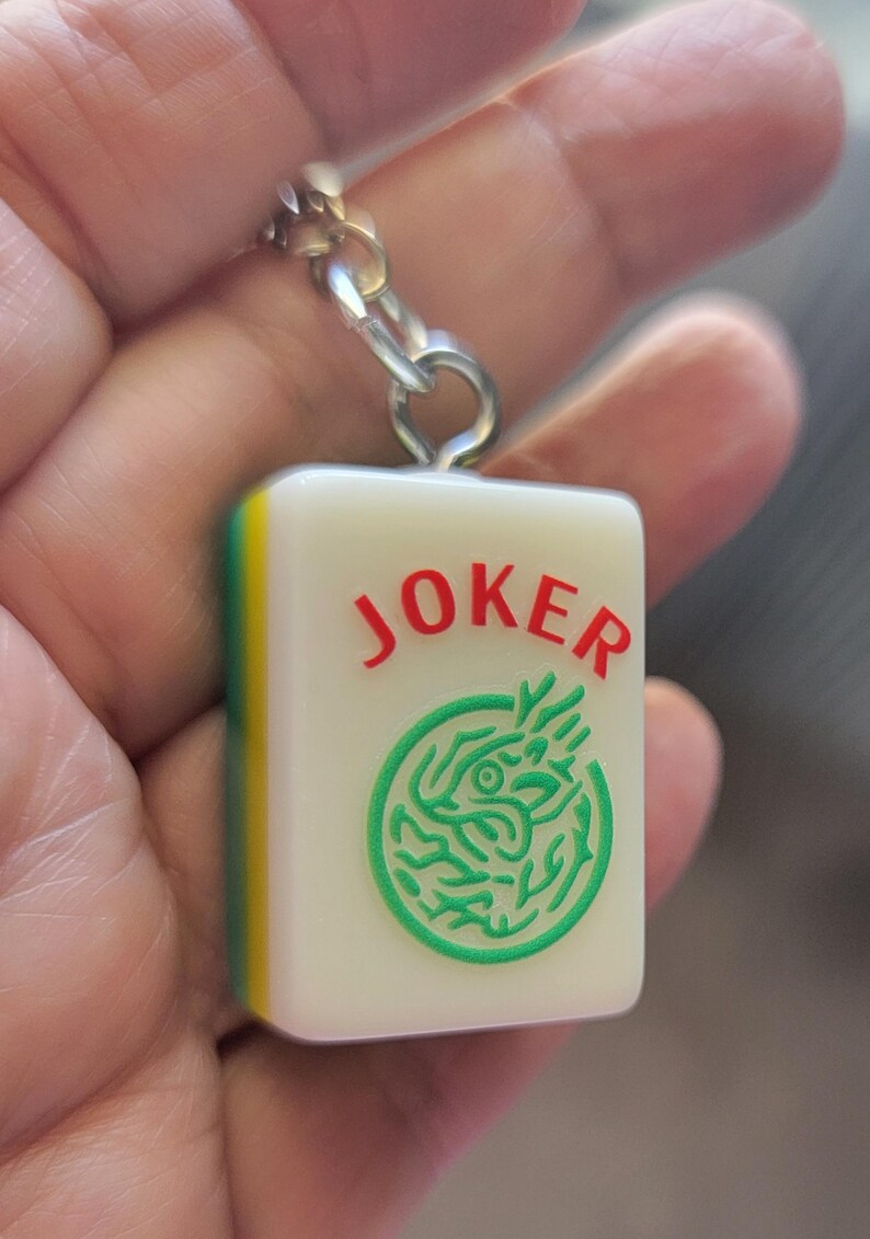 May include: A white Mahjong tile keychain with the word "JOKER" in red and a green dragon design. The tile has a yellow and green edge and is attached to a silver chain.