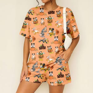 May include: Orange Halloween-themed pajama set featuring a repeating pattern of cartoon characters, pumpkins, ghosts, and castles. The set includes a short-sleeved t-shirt and matching shorts. A white bag is worn over the shoulder.