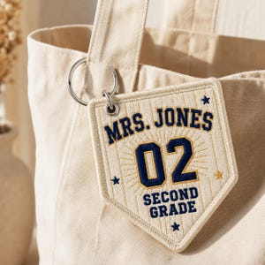 May include: A cream-coloured canvas tote bag featuring a pentagonal embroidered patch attached with a silver metal keyring. The patch displays the text MRS. JONES, the number 02, and SECOND GRADE in navy blue, accented with small embroidered stars.