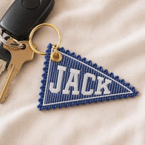 Könnte beinhalten: Ein dreieckiger Schlüsselanhänger aus blau-weiß gestreiftem Stoff mit gezackten Rändern, auf dem der Name JACK in weißen Blockbuchstaben gestickt ist. Der Stoffwimpel ist an einem goldfarbenen Metallring befestigt, der einen schwarzen Autoschlüssel hält.