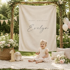 May include: A beige fabric backdrop featuring the text HAPPY BIRTHDAY and the name Evelyn in black lettering, suspended from a wooden frame. A child sits on a textured blanket near a wicker basket, a plush rabbit, and floral arrangements.