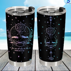 Puede incluir: Dos tazas de viaje de acero inoxidable negro con un diseño celestial. El diseño incluye una luna creciente, manos, estrellas y el texto "You are not alone in this - NONE OF US ARE -". Las tazas tienen una base plateada y una tapa negra.
