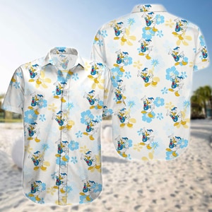Può includere: Camicia bianca a maniche corte con un motivo ripetuto di personaggi dei cartoni animati di Paperino e accenti floreali blu e gialli. La camicia è esposta su uno sfondo di spiaggia.