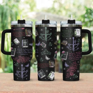 May include: Three black insulated travel mugs with handles, each adorned with fantasy-themed illustrations. The designs include books, butterflies, swords, and floral elements in various colours. Each mug has a black lid and straw. Text is also present.