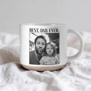 May include: White ceramic mug with a beige base and a curved handle. The mug features a black and white photo of a man and a child, with the text "BEST. DAD. EVER." printed above the photo.