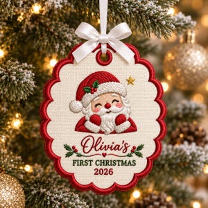 May include: An oval-shaped embroidered Christmas ornament with a scalloped red border and a white satin ribbon bow. The centre features a smiling Santa Claus, the text "Olivia's FIRST CHRISTMAS 2026," and small green holly sprigs with red berries.