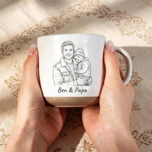 May include: White ceramic mug with a brown base, featuring a black and white illustration of a father holding a child. The text "Ben & Papa" is printed below the illustration. The mug has a curved handle.