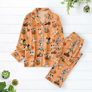 May include: Orange Halloween pyjama set with a button-down top and matching trousers. The set features a repeating pattern of cartoon characters, pumpkins, and haunted houses. White trim accents the collar, cuffs, and pockets.