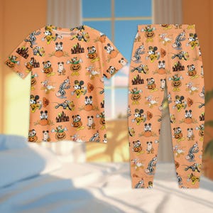 May include: Orange Halloween-themed pajama set featuring a short-sleeved top and matching pants. The set is covered in a repeating pattern of Mickey Mouse, pumpkins, ghosts, and haunted houses. The pajamas are made of a soft, comfortable material.
