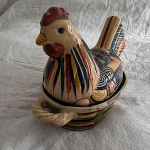 May include: A hand-painted ceramic chicken-shaped container. The chicken has a red comb, blue eye, and a yellow beak. The body is decorated with vertical stripes in various colors. The container has a handle and is designed to hold items.