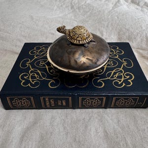 May include: A decorative, antique-style trinket box with a turtle figurine on top. The box is dark metallic with a white rim, and sits on a dark blue book with gold filigree and the title "Wuthering Heights" by Emily Brontë.