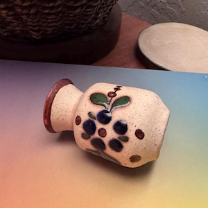 May include: A small, white ceramic vase with a brown rim and a painted floral design. The vase features a blue flower with green leaves and brown dots.