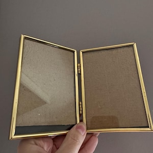 May include: A gold-colored, hinged double picture frame. The frame is open, revealing two empty spaces for photos. The frame has a simple, elegant design with a thin border. The background is a solid gray color.