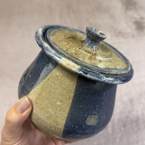May include: A handmade ceramic jar with a lid. The jar is a rounded shape with a blue and tan color scheme. The lid has a small knob and a blue rim. The jar is held by a person.