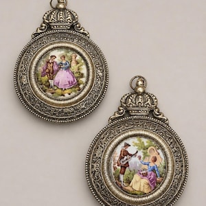 May include: Two circular metal pendants with an antique brass finish and crown-shaped bails. Each pendant features a central porcelain inlay depicting a colorful scene of two figures in period clothing within a garden setting, framed by ornate metalwork.