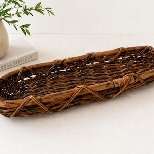May include: A long, oval-shaped woven basket in shades of brown. The basket is made of natural materials, with a textured surface and a decorative crisscross pattern around the edges. The basket is empty and sits on a white surface.