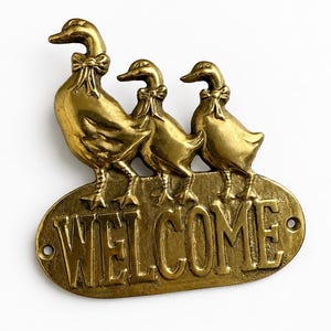 May include: A brass-colored welcome sign featuring three ducks with bow ties. The ducks stand on an oval plaque with the word "WELCOME" in embossed lettering. The sign is designed for indoor or outdoor use.