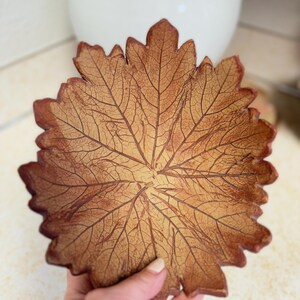 May include: A decorative leaf-shaped bowl with a detailed vein pattern. The bowl is a warm brown color with darker brown edges, resembling a fall leaf. The bowl is held in a hand, suggesting its size and use as a decorative item.