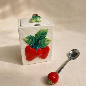 May include: A white ceramic sugar bowl with a square shape and a lid, decorated with a 3D strawberry design. A small stainless steel spoon with a red strawberry-shaped handle is next to the bowl. The bowl has a textured pattern.