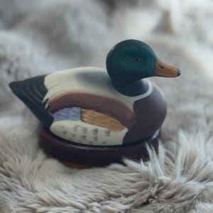 May include: A hand-painted wooden duck decoy with a green head, brown body, and white markings. The duck is sitting on a wooden base.