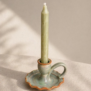May include: A handmade ceramic candlestick holder with a green glaze and a brown rim. A long, green candle is inserted in the holder. The candlestick has a handle and a scalloped base. The background is a neutral color.