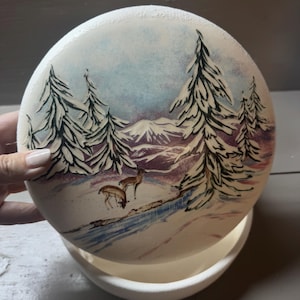 May include: A round, white ceramic planter with a hand-painted winter scene. The scene depicts snow-covered pine trees, mountains, and two deer near a stream. The planter has a matching saucer.
