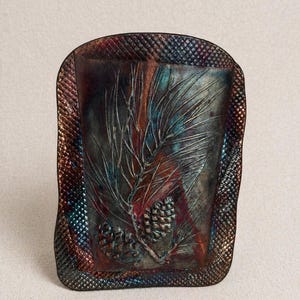 May include: A rectangular, metallic tray with a textured border. The center features an embossed design of pine needles and cones. The color palette includes shades of blue, red, and brown, creating a rustic, artistic aesthetic.