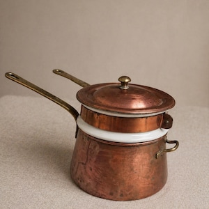 May include: A copper double boiler with a lid and brass handles. The pot has a white ceramic insert and a small brass knob on the lid. The copper has a warm, reddish-brown hue, and the overall design is vintage.
