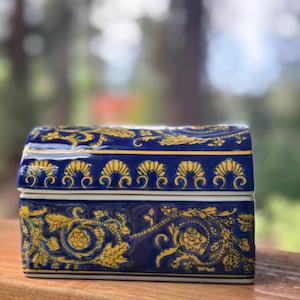 May include: A rectangular, blue ceramic trinket box with a hinged lid. The box is decorated with gold floral and scrollwork patterns. The box sits on a wooden surface, with a blurred background of green foliage.