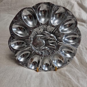 May include: A silver-colored egg serving platter with a bunny design in the center. The platter has 12 egg-shaped indentations around the edge. The platter is made of metal and has a shiny finish.