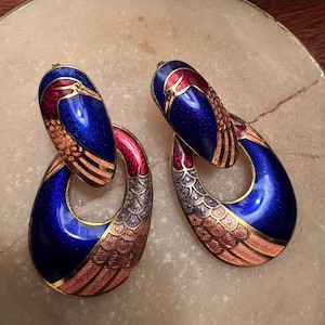 May include: A pair of vintage earrings with a bird design. The earrings feature a blue, red, and gold color scheme. The bird's head is at the top, with a teardrop-shaped body below. The earrings are made of metal and enamel.