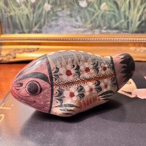 May include: A hand-painted ceramic fish figurine. The fish is primarily pink with black, white, and red decorative patterns. The fish has a large eye and a black tail. The figurine is resting on a dark surface.