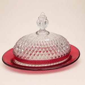 May include: A clear glass butter dish with a diamond pattern dome and a ruby red base. The dish has a decorative finial on top of the dome. The base is a matching ruby red color.
