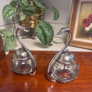 May include: Two vintage silver-plated swan-shaped condiment jars. Each jar has a clear glass container with a lid and a swan-shaped handle. The jars are displayed on a wooden surface, with a painting and a potted plant in the background.