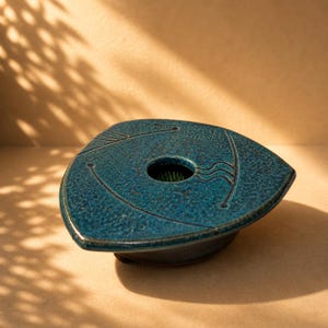 May include: A teal ceramic flower frog with a textured surface and a central hole for arranging flowers. The triangular shape features etched lines and a dark rim. The frog is designed for floral arrangements.