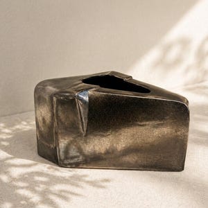 May include: A geometric, metallic vase with a dark, textured finish. The vase has an angular shape with a rectangular base and a slanted top opening. The color is a mix of bronze and black, with visible crackling on the surface.