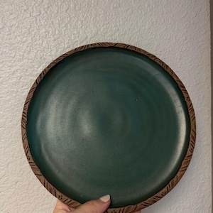 May include: A handmade ceramic plate with a deep teal center and a textured, brown rim. The plate is round and appears to be stoneware. The rim has a repeating pattern. The plate is held by a hand.