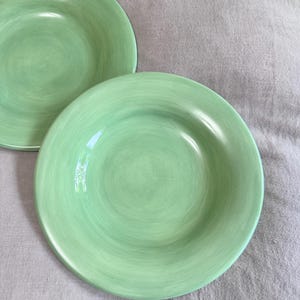 May include: Two light green ceramic plates with a slightly textured surface. The plates have a shallow bowl shape and a wide rim. The plates are a solid, pale green color and are set against a neutral background.