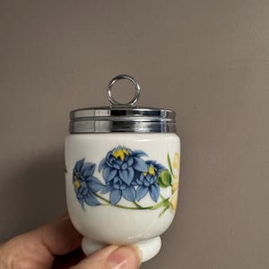 May include: A white porcelain egg cup with a floral design featuring blue and yellow flowers. The egg cup has a silver-colored metal lid with a ring handle. The design is a classic style, suitable for serving boiled eggs.
