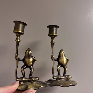 May include: A pair of brass frog candle holders. Each frog figure holds a candle cup. The frogs stand on a leaf-shaped base. The candle holders have a vintage, antique appearance.