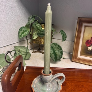 May include: A tall, pale green candle stands in a handmade ceramic candle holder. The candle holder is light green with a scalloped edge and a small handle. The candle holder sits on a wooden surface.