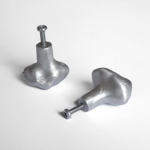 Op de afbeelding: Twee zilverkleurige kastknoppen met een getextureerde, organische vorm. Elke knop heeft een schroefdraad voor bevestiging. De knoppen worden op een witte achtergrond getoond, wat hun metallic afwerking en ontwerp benadrukt.