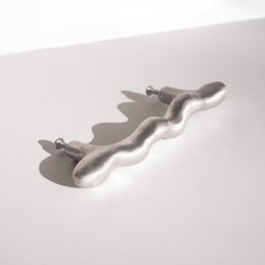 以下が含まれることがあります： シルバーカラーの、波打つキャビネットプル。2つの取り付けポストが付いています。この金具は滑らかで有機的な形状をしており、引き出しやキャビネットでの使用を想定しています。白い背景に影を落として表示されています。
