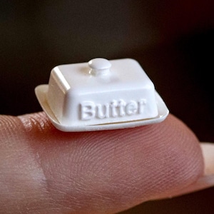Könnte beinhalten: Eine Miniatur-Butterdose aus weißem Kunststoff mit einem Deckel, der einen kleinen Knaufgriff besitzt. Das Wort Butter ist in Großbuchstaben auf der Vorderseite des Deckels eingeprägt. Das Objekt ist klein genug, um auf einer Fingerspitze zu balancieren.