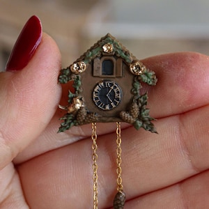 Pode incluir: Relógio de cuco em miniatura com moldura de madeira castanha, detalhes de folhagem verde e enfeites dourados. O mostrador do relógio é preto com algarismos romanos brancos. Duas correntes douradas pendem com pequenos pesos de pinha.