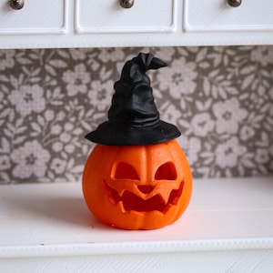 Peut inclure: Une figurine de citrouille jack-o'-lantern orange vif présentant un visage sculpté avec des yeux triangulaires et une bouche dentelée, surmontée d'un petit chapeau de sorcière noir texturé posé sur la surface arrondie de la citrouille.