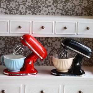 Puede incluir: Dos batidoras de pie en miniatura con varillas metálicas plateadas. Una batidora es de color rojo brillante con un cuenco estriado azul claro, y la otra es negra brillante con un cuenco estriado beige. Ambas tienen detalles plateados.