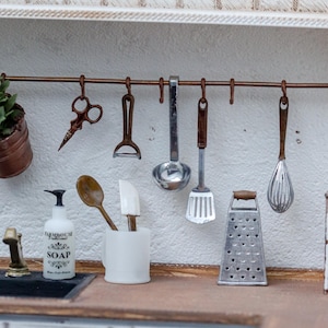 Könnte beinhalten: Eine Miniatur-Küchenszene mit einer kupferfarbenen Hängestange und kleinen Metall-Küchenutensilien wie Schere, Sparschäler, Schöpfkelle, Pfannenwender und Schneebesen. Ein weißer Seifenspender mit der Aufschrift FARMHOUSE SOAP, ein Holzlöffel und eine Reibe.