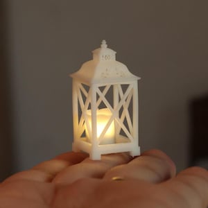 Könnte beinhalten: Eine weiße Miniatur-Laterne mit quadratischem Sockel und kreuzförmigen Streben an den Seiten, in der ein kleines, warm leuchtendes LED-Licht brennt. Die Laterne hat ein verziertes Oberteil mit Ausschnitten und einer spitzen Verzierung.