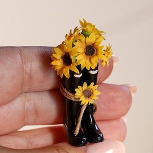 Puede incluir: Un broche decorativo en miniatura que presenta un par de botas de lluvia negras brillantes llenas de girasoles amarillos. Un pequeño girasol está atado al frente de las botas con un cordel marrón fino, y cada bota tiene un pequeño detalle blanco.